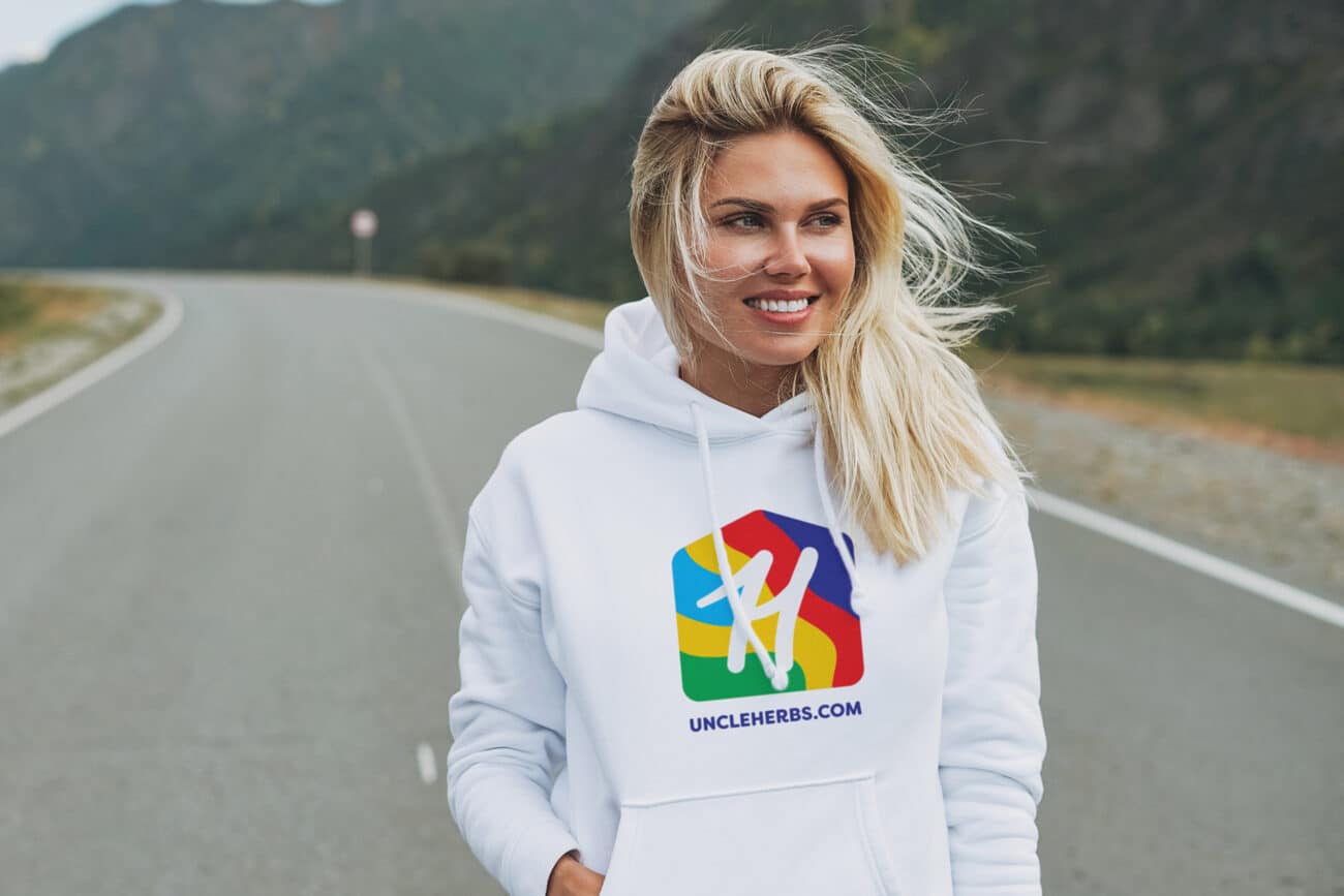 Beautiful smiling blonde young woman traveler in white hoodie with Uncle Herb's Dispensary Logo on road, trip to mountains,